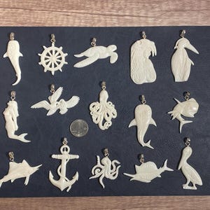 May include: A collection of 16 white bone pendants featuring various sea life designs, including a mermaid, octopus, fish, turtle, and anchor. The pendants are displayed on a black background.