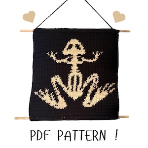 May include: A black crochet wall hanging featuring a cream-colored frog skeleton design. The wall hanging is suspended by a black string and wooden dowels. The text "PDF PATTERN !" is displayed at the bottom.