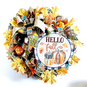 以下が含まれることがあります： カボチャが描かれた「Hello Fall」のサインが付いた、丸い秋のリース。リースには、人工の紅葉、ヒョウ柄のリボン、小さなカボチャの装飾が含まれています。カラーパレットには、オレンジ、ブラウン、グリーン、イエローが含まれています。
