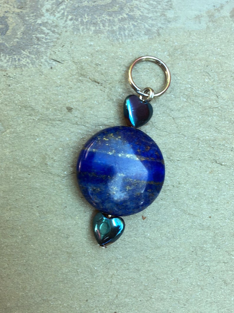 May include: A blue lapis lazuli stone pendant with a silver ring at the top and two iridescent blue heart-shaped beads.