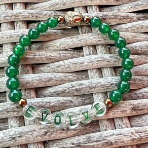 May include: A bracelet with green beads and clear beads spelling "EVOLVE". The bracelet has a gold-coloured Buddha head and gold-coloured accent beads. The bracelet is set against a woven background.