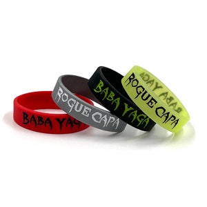 May include: Four rubber wristbands in red, gray, black, and neon green. Each band has the words "BABA YAGA" or "ROGUE CAPA" in a stylized font. The bands are stacked slightly, showing the text on each.