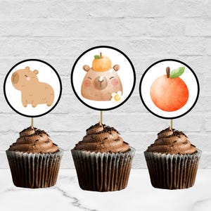 Peut inclure: Trois cupcakes au chocolat avec des décorations. Les décorations représentent un capybara de dessin animé, un ours avec une orange sur la tête et une orange à l'aquarelle. Les cupcakes sont dans des caissettes marron et saupoudrés de vermicelles au chocolat.