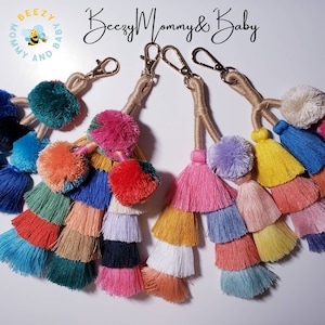 May include: Set of five colorful tassel keychains with pom poms. Each keychain has a gold metal clasp and is made of multiple layers of colorful tassels. The keychains are arranged in a semi-circle on a white surface. The text "Beezy Mommy & Baby" is visible in the background.