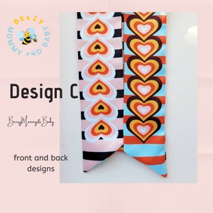 May include: Two fabric hair scrunchies with a heart pattern. The scrunchies are pink and blue with black and orange stripes. The scrunchies are shown front and back. The text "Beezy Mommy & Baby" is printed on the scrunchies.