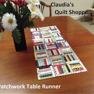 Könnte beinhalten: Patchwork-Tischläufer mit einer Vielzahl von bunten Stoffen in einem geometrischen Muster. Der Läufer liegt auf einem Holztisch mit einer Blumenvase im Hintergrund. Der Text "Claudia's Quilt Shoppe" ist im Bild sichtbar.