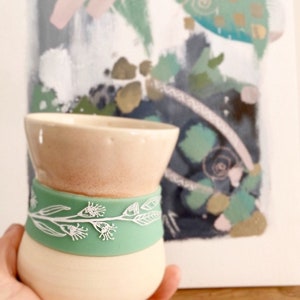 May include: A handcrafted ceramic cup with a beige top, a pale green band featuring white floral patterns, and a cream-coloured base. The cup is held in a hand, with an abstract painting in the background.