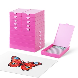 Może przedstawiać: Stos różowych pudełek do przechowywania i otwarte pudełko ze srebrnymi koralikami. Ukończony haft krzyżykowy przedstawiający motyla z czerwonymi, pomarańczowymi, niebieskimi i czarnymi detalami spoczywa na białej powierzchni.