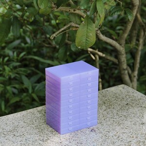 May include: A stack of twelve rectangular, translucent light purple plastic storage cases. Each individual case features a hinged lid with a small tab closure, arranged in two vertical columns of six units each, creating a uniform block structure.
