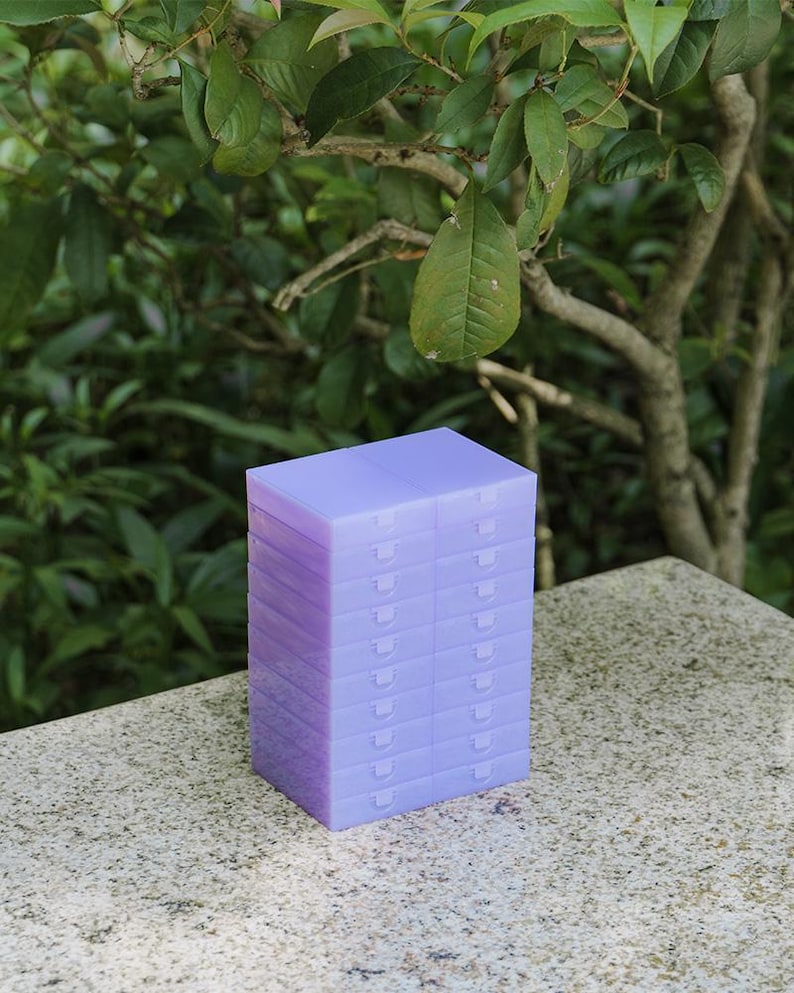 May include: A stack of twelve rectangular, translucent light purple plastic storage cases. Each individual case features a hinged lid with a small tab closure, arranged in two vertical columns of six units each, creating a uniform block structure.