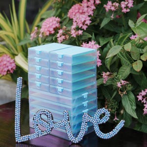 May include: A stack of clear, light blue plastic storage boxes with matching lids. The word "love" is spelled out in a decorative script, embellished with small, iridescent beads. Pink flowers and green foliage are in the background.