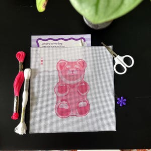 May include: A needlepoint canvas featuring a pink gummy bear design, accompanied by a skein of pink embroidery floss, a skein of white embroidery floss, a pair of small white-handled scissors, and a small purple flower-shaped needle minder.