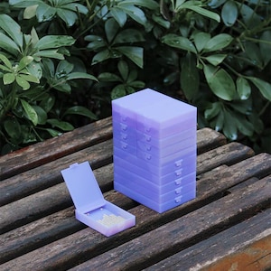 May include: A stack of eight rectangular light purple plastic storage cases with hinged lids, accompanied by one identical open case lying flat in front, revealing a small collection of tiny white beads stored inside the translucent container.