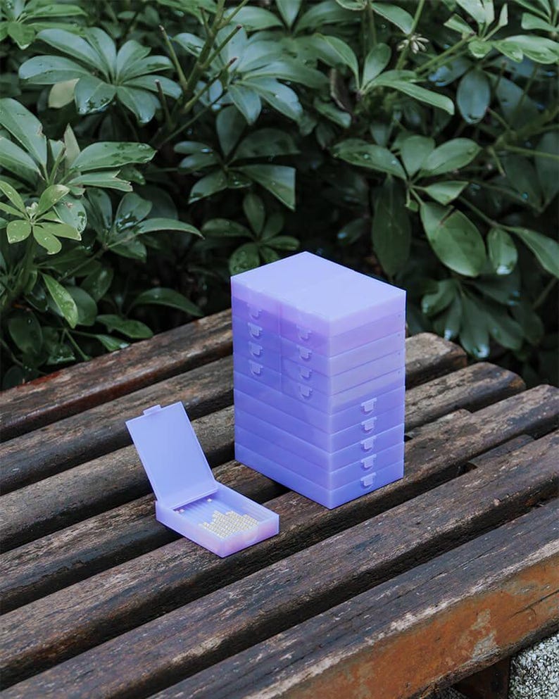 May include: A stack of eight rectangular light purple plastic storage cases with hinged lids, accompanied by one identical open case lying flat in front, revealing a small collection of tiny white beads stored inside the translucent container.