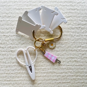 Könnte beinhalten: Ein goldfarbener Metallkarabiner-Schlüsselanhänger mit mehreren weißen Kunststoff-Garnspulen, einer Stickschere mit weißen Kunststoffgriffen, einem kleinen Metall-Nahttrenner mit einem rosa-weiß karierten Stoffgriff und einem Gänseblümchen-Anhänger.