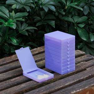 May include: A stack of nine rectangular light purple plastic storage cases with hinged lids, positioned next to a single open case of the same design containing a small collection of thin, light-coloured cylindrical sticks inside the base.