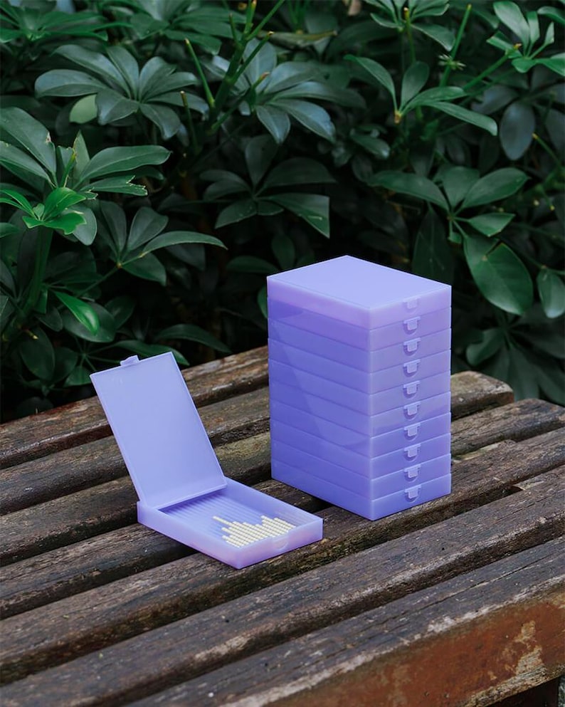 May include: A stack of nine rectangular light purple plastic storage cases with hinged lids, positioned next to a single open case of the same design containing a small collection of thin, light-coloured cylindrical sticks inside the base.