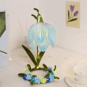 May include: A decorative flower-shaped lamp with a light blue, translucent shade. The lamp has a green stem and leaves, and is surrounded by a small wreath of blue and yellow flowers. The lamp is on a white surface.