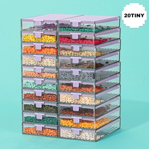 May include: A stack of clear plastic storage containers with lavender lids and latches. Each container is filled with small, colorful craft supplies. The text "20TINY" is in a white square in the upper right corner.