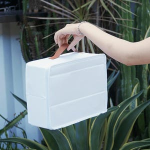 May include: A rectangular white storage case with a smooth, solid-coloured exterior and a brown leather-like handle attached to the top. The case features a structured, boxy design with clean lines and a minimalist aesthetic for organising items.