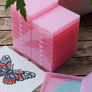 May include: Pink plastic storage boxes stacked on a wooden surface, with a butterfly diamond painting and a tray of blue beads. The boxes are square and have a hinged lid. The butterfly design is in shades of gray, orange, and black.