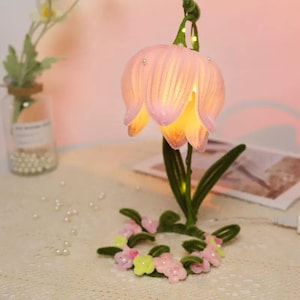 May include: A decorative flower-shaped lamp with a pink, translucent shade and a warm yellow glow. The lamp has green leaves and a base adorned with small pink and yellow flowers. The lamp is on a table with a glass jar and pearls.