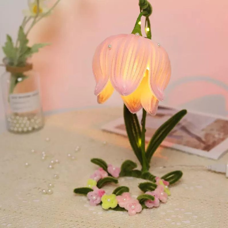 May include: A decorative flower-shaped lamp with a pink, translucent shade and a warm yellow glow. The lamp has green leaves and a base adorned with small pink and yellow flowers. The lamp is on a table with a glass jar and pearls.