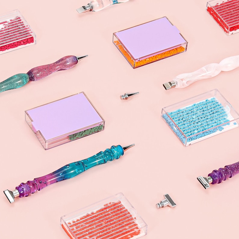 May include: An assortment of diamond painting tools and accessories are arranged on a light pink surface. The tools include pens with colorful, glittery handles, small trays filled with colorful beads, and metal applicator tips. The items are for crafting and art projects.