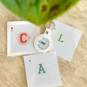 May include: Three square white needlepoint canvases featuring stitched letters C, L, and A in red and green thread, accompanied by a white circular leather keychain holder with a metal ring and a 3M adhesive backing label.