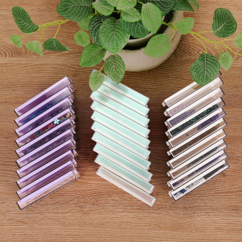 May include: An overhead shot of three stacks of rectangular, clear plastic cases. The cases are angled and contain various items, including glitter and floral designs. The cases are arranged on a wooden surface, with a potted plant in the background.
