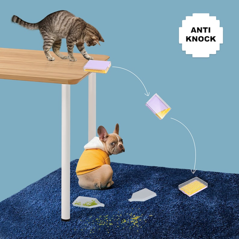 May include: A tabby cat on a wooden table knocks over a container of yellow beads, with a French bulldog in a yellow hoodie watching. The text "ANTI KNOCK" is in a white square. The beads are spilling onto a blue rug.