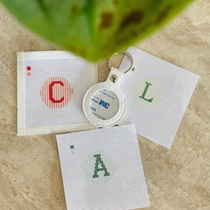 May include: Three square white needlepoint canvases featuring cross-stitched letters C, L, and A in red and green, accompanied by a white circular leather keychain holder with a metal ring and a central 3M adhesive backing label.