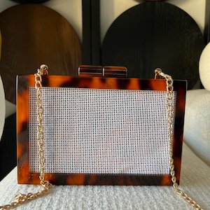 May include: A rectangular box clutch with a brown and black tortoiseshell pattern frame and a white grid mesh center panel. The bag features a gold-tone metal chain strap and a matching tortoiseshell clasp closure on the top edge of the frame.