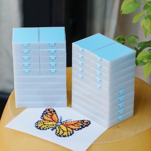 May include: Several clear plastic craft storage boxes with light blue lids and clasps. A cross-stitch butterfly with orange, yellow, and black details is on white paper. The boxes are on a wooden surface.