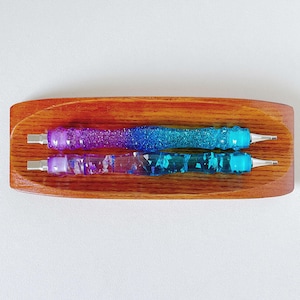 May include: Two sparkly pens with a gradient of purple, blue, and turquoise colors. The pens are in a wooden holder.