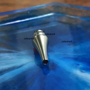 May include: A silver metal nozzle tip for an airbrush. The tip is pointed and has a small hole at the end.