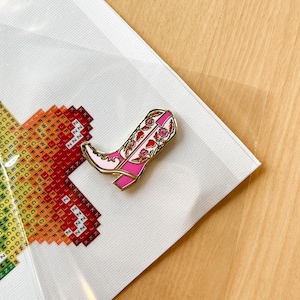 May include: A pink and white cowboy boot with floral embroidery, gold accents, and a white background. The boot is a pin.