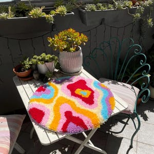 Op de afbeelding: Een rond, kleurrijk tapijt met een draaiende print in tinten roze, geel, blauw en rood. Het tapijt ligt op een witte tafel met een groene metalen stoel op de achtergrond.