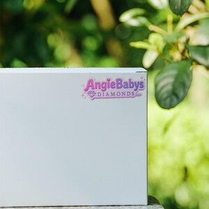 May include: A rectangular white box featuring a sticker with the purple text AngieBabys DIAMONDS and a small diamond icon. The sticker includes decorative star accents and a thin purple border, positioned on the upper right side of the box.