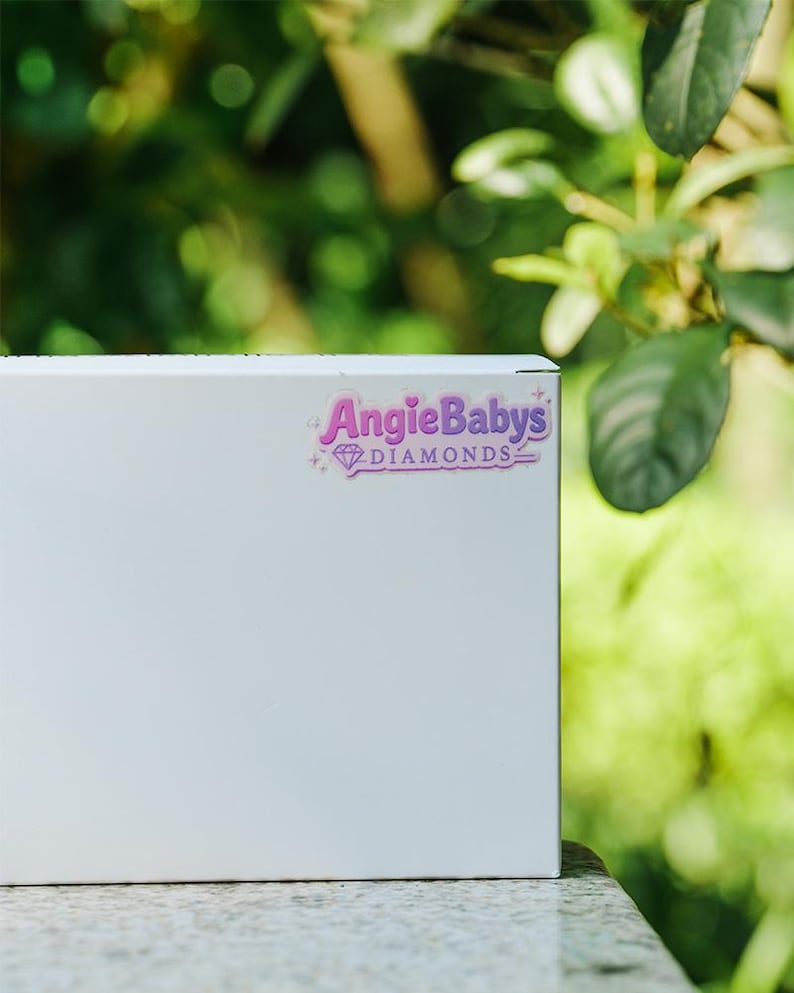 May include: A rectangular white box featuring a sticker with the purple text AngieBabys DIAMONDS and a small diamond icon. The sticker includes decorative star accents and a thin purple border, positioned on the upper right side of the box.