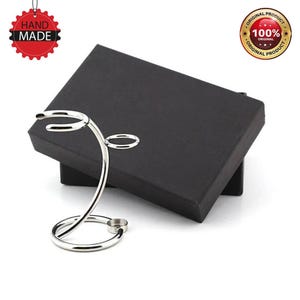 Puede incluir: Un soporte de afeitar de metal plateado con un diseño curvo. El soporte está sobre una caja negra con las palabras "Original Product 100% Original Product" impresas en un círculo blanco.
