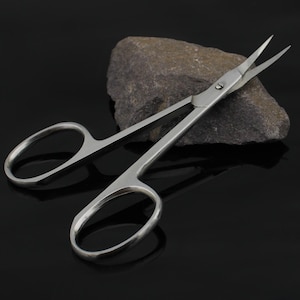 May include: A pair of silver-toned cuticle scissors with a curved blade and a pointed tip. The scissors are lying on a gray rock.