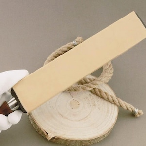 May include: A beige leather strop with a wooden handle, used for sharpening knives. The strop is held over a wooden slice and a coiled rope, suggesting a rustic or artisanal theme. The handle is dark brown.