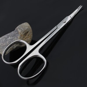 May include: A pair of stainless steel cuticle scissors with sharp pointed tips. The scissors are closed and lying on a black surface.
