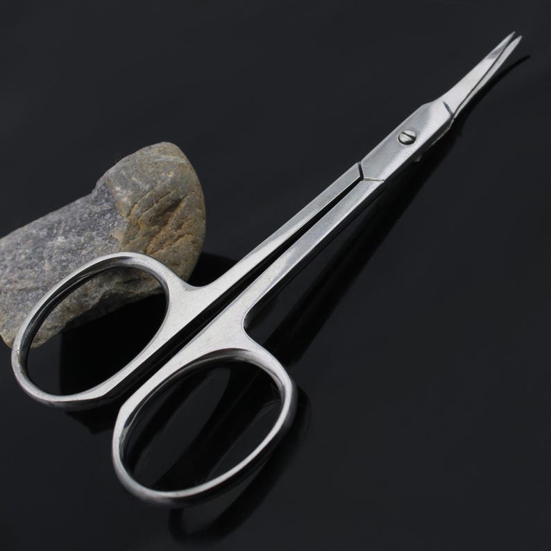 May include: A pair of stainless steel cuticle scissors with sharp pointed tips. The scissors are closed and lying on a black surface.