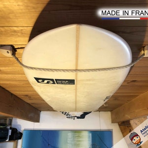 May include: A white surfboard with a rope attached to it, hanging from the ceiling of a wooden room. The surfboard has a black logo that says "SC.N. SURFBOARDS" and the text "MADE IN FRANCE" is visible on the ceiling.