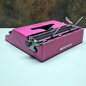 May include: A pink vintage typewriter with a black roller and silver keys. The typewriter is closed and the carriage return lever is raised.