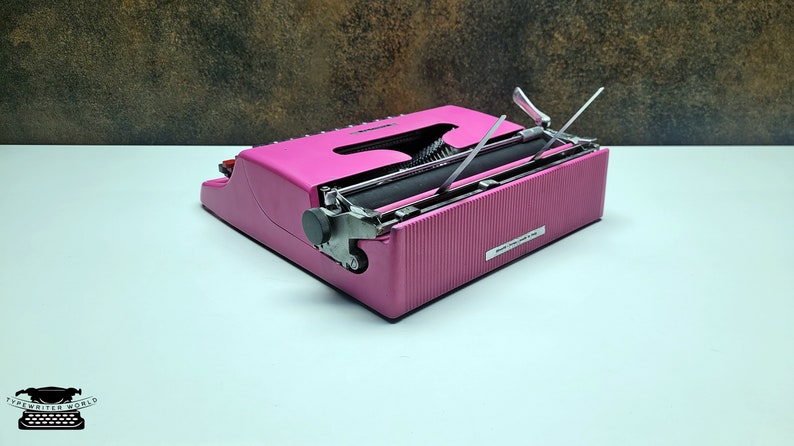 May include: A pink vintage typewriter with a black roller and silver keys. The typewriter is closed and the carriage return lever is raised.