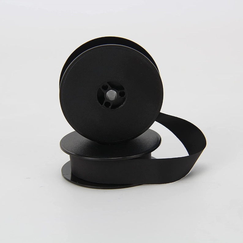 May include: Two black typewriter ribbon spools, one on top of the other. The ribbon is wound around the spool and is ready to be used in a typewriter.