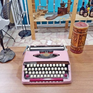 May include: A pink Brother typewriter with a white top and a brown wooden base. The typewriter has a standard keyboard layout with keys labeled in English. The typewriter is sitting on a wooden table.